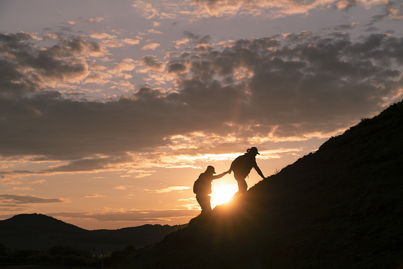 在多云的日落背景下，人們幫助朋友爬山的剪影圖片素材