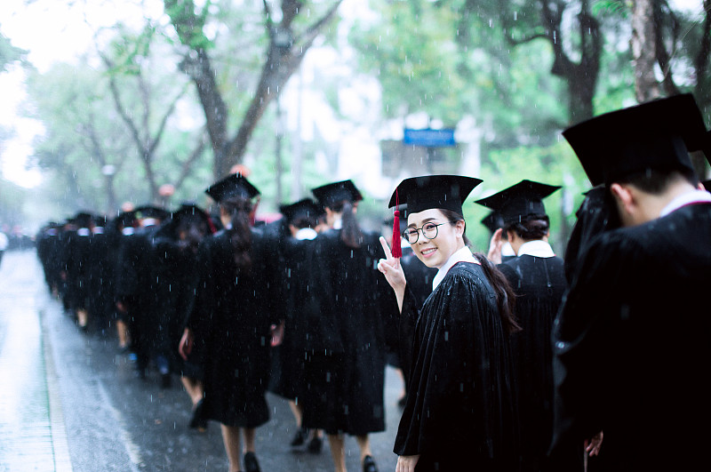 一個快樂的年輕女子戴著學士帽，在雨中與朋友們在路上做著和平手勢圖片素材