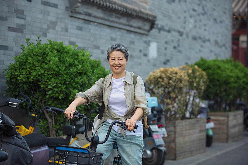 老年人City walk系列:老年女人骑共享单车在北京胡同旅行图片下载