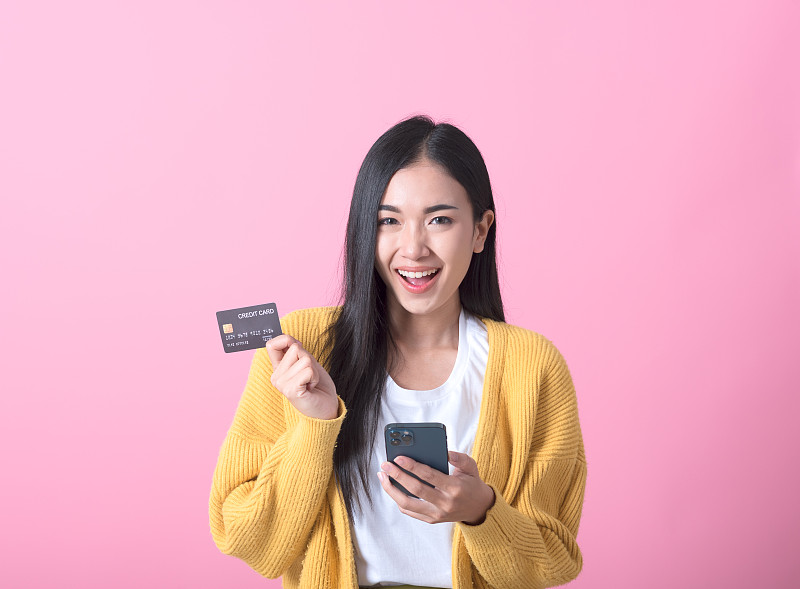 一個快樂的年輕女孩的肖像顯示塑料信用卡持有手機隔離在粉紅色的背景圖片素材