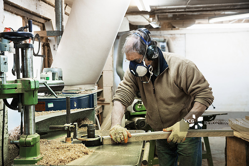 一名男子站在木工车间里，戴着呼吸器和听力保护器，在一块木头上工作。图片素材