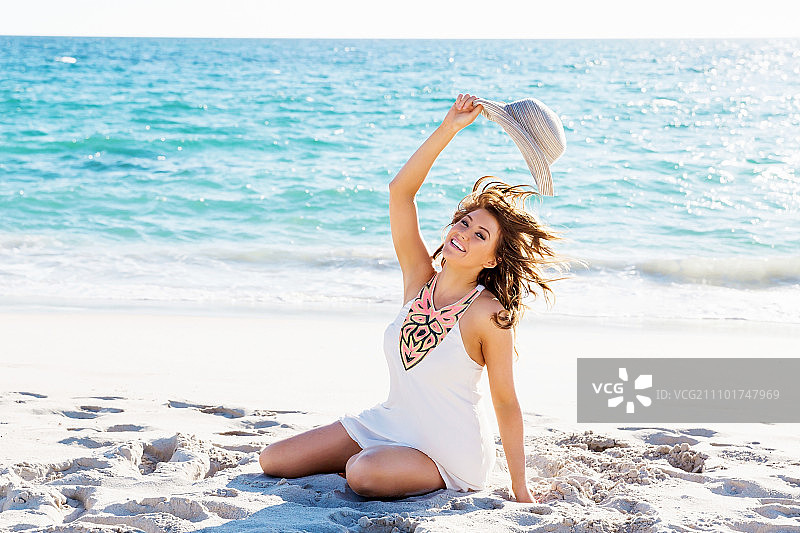 坐在海滩上的年轻女子。一个戴着帽子坐在海滩上的年轻漂亮女人的肖像图片素材