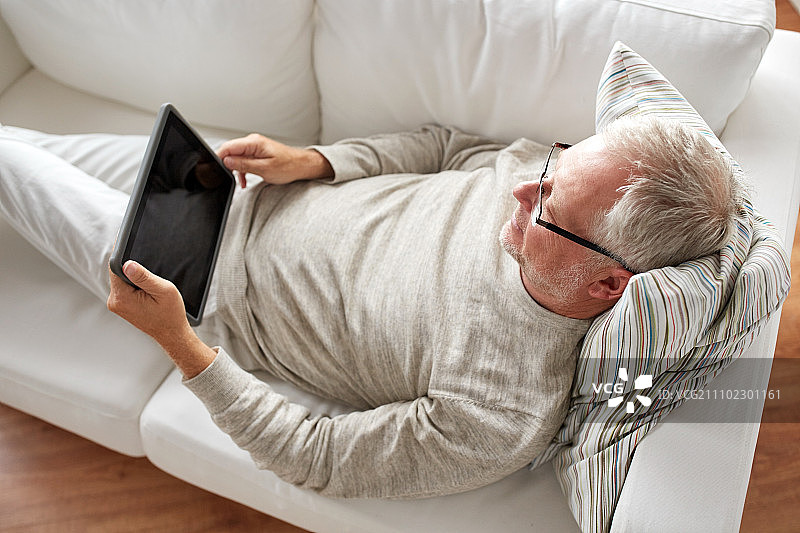 技术、人与生活理念——老年人用黑色的空白平板电脑屏幕躺在家里的沙发上。老人拿着平板电脑躺在家里的沙发上图片素材