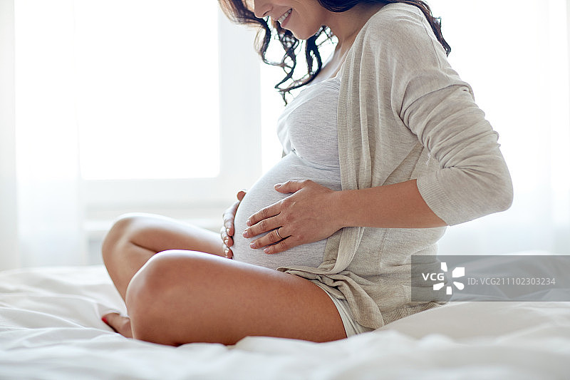 怀孕，休息，人们和期望的概念-近距离的快乐微笑孕妇坐在床上，触摸她的肚子在家里图片素材