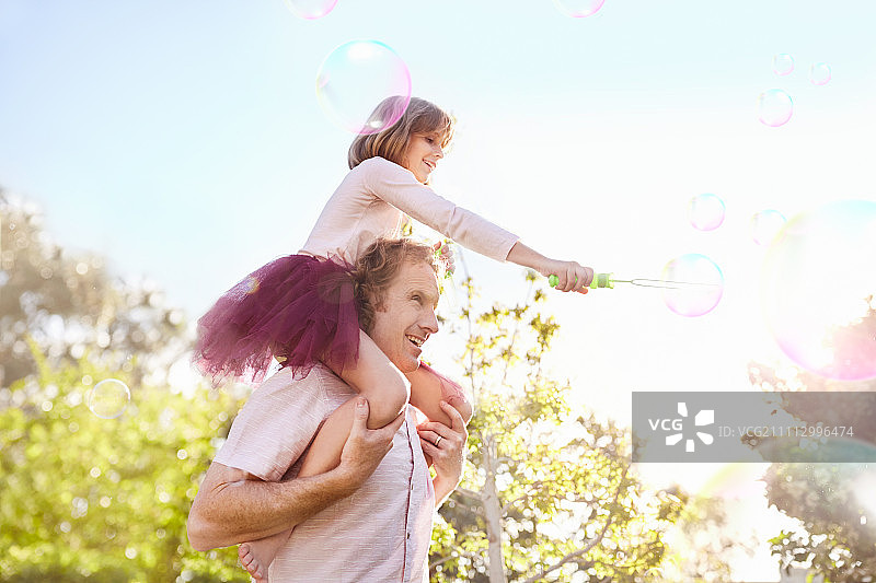 在阳光明媚的夏日公园里，父亲扛着肩上带着泡泡棒的女儿图片素材