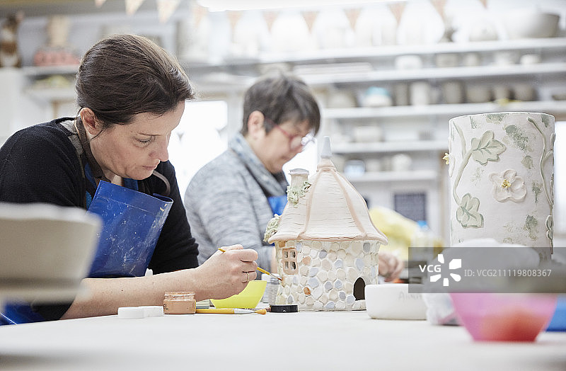 两名妇女坐在一个陶器工作室的工作台前，制作装饰的陶土制品。图片素材