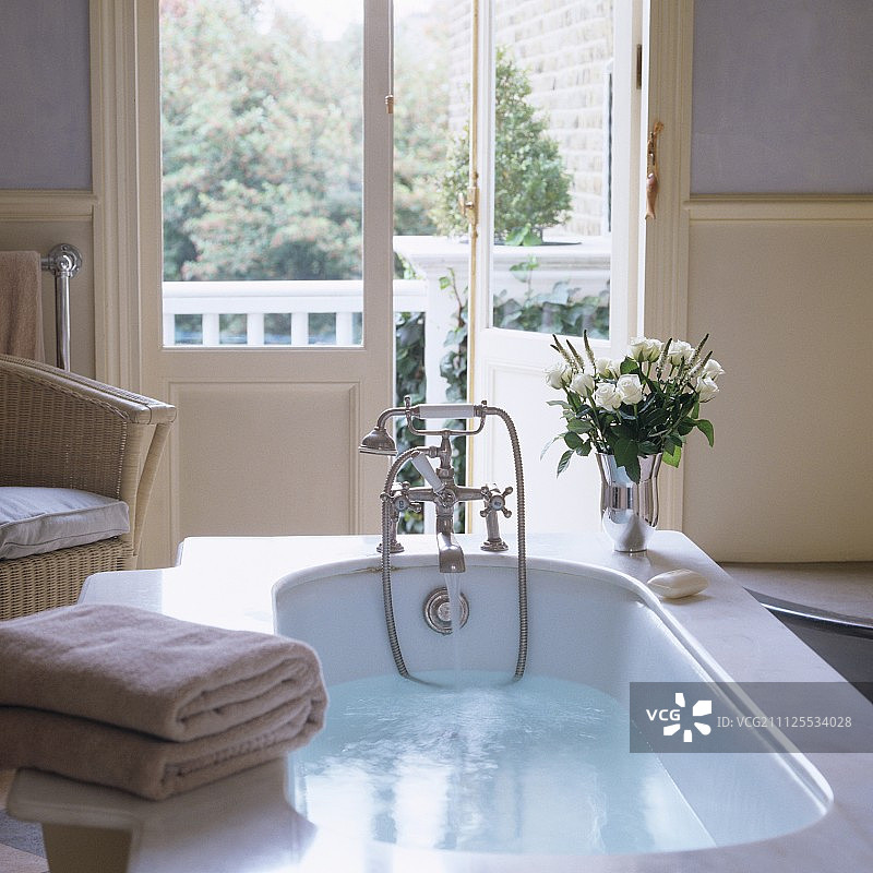 浴缸中的洗澡水与瓶白玫瑰在浴缸周围，透过半开的法式门进入花园图片素材