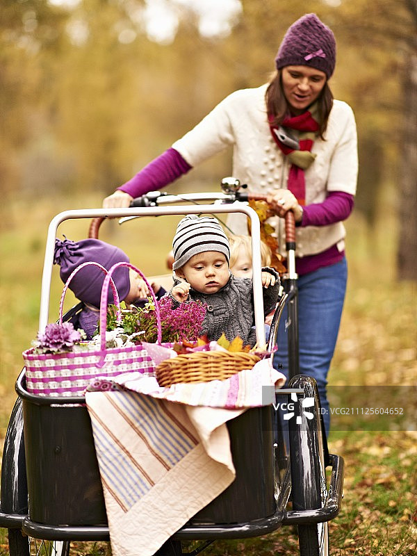 母亲推着孩子们和野餐篮在箱子的货物自行车通过秋天的风景图片素材