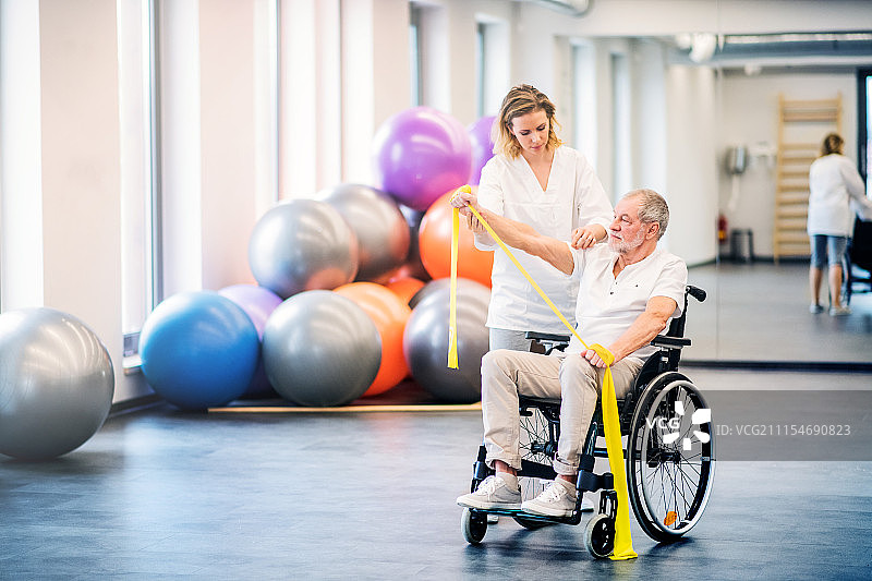 年轻女性理疗师与一位坐轮椅的老年男子一起工作。图片素材