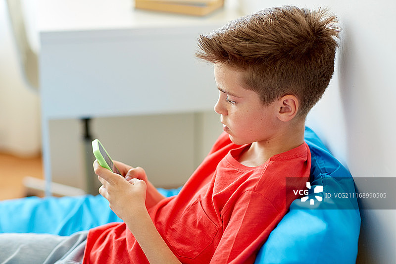 童年时代，网络欺凌和交流概念——男孩躺在家里的床上玩智能手机。男孩躺在家里的床上拿着智能手机图片素材