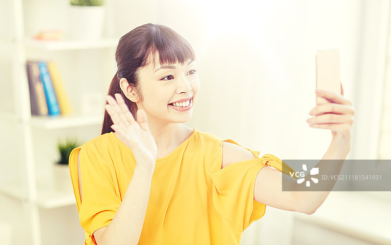 人、科技和休闲概念——快乐的年轻亚洲女性在家里用智能手机自拍并挥手。快乐的亚洲女人用智能手机自拍。快乐的亚洲女人用智能手机自拍图片素材