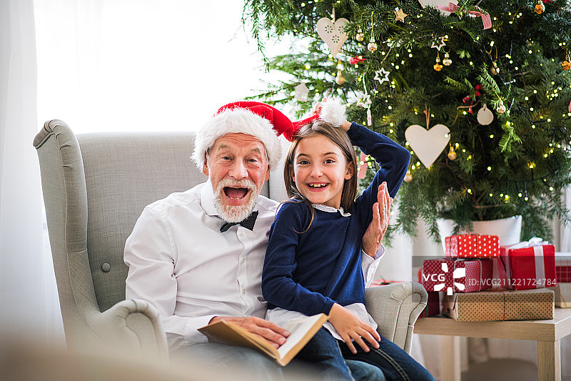 一个小女孩和她的爷爷带着圣诞帽和一本书在圣诞节。图片素材