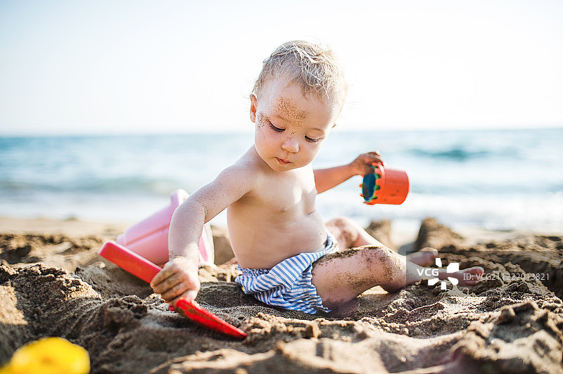 暑假，一个蹒跚学步的小女孩坐在海滩上玩耍。图片素材