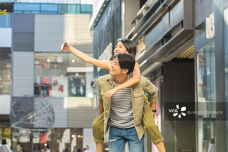 青年男子背着青年女子在户外商业街旅游用手机自拍嬉戏玩耍图片素材