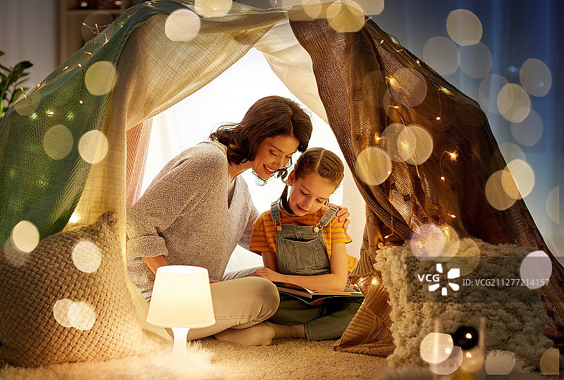 家庭、舒适和人的概念——快乐的母亲和小女儿晚上在家里的孩子帐篷里看书。快乐的家庭在家里的孩子帐篷里看书图片素材