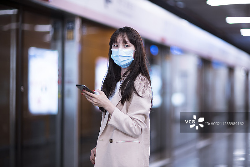 年轻女子戴着防护口罩等候地铁并使用智能手机图片素材
