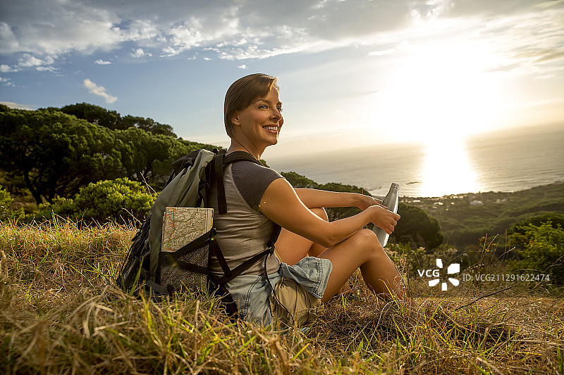 微笑的女子背包坐在草地上的海岸景观在日落图片素材