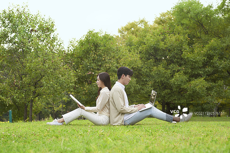 一对夫妇背靠背坐在草地上看书，看着笔记本电脑的照片图片素材
