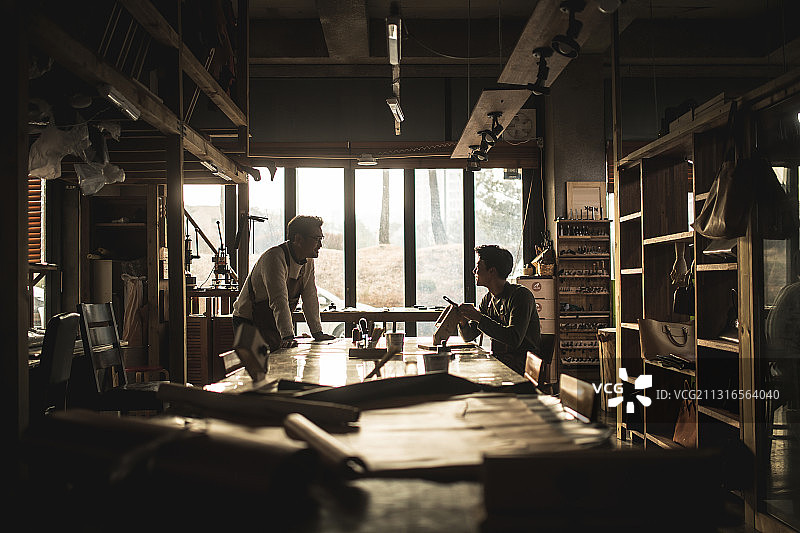 一个成熟的男人和一个年轻人坐在车间的桌子旁谈论着皮革工艺图片素材