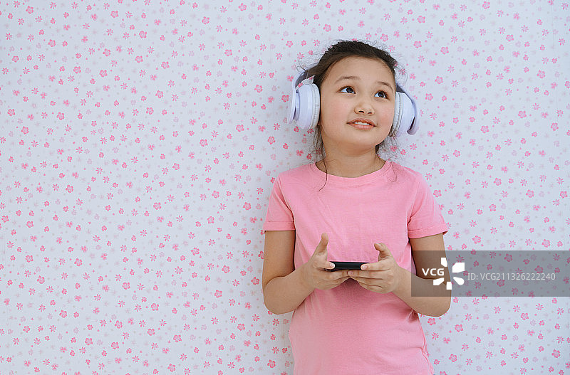 可爱的女孩戴着耳机靠在粉红色的墙上玩着智能手机图片素材