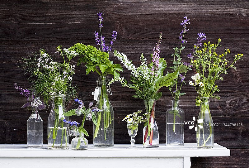在深色木墙的衬托下，白色的桌子上，玻璃瓶和玻璃瓶里装着盛开的花园草本植物图片素材