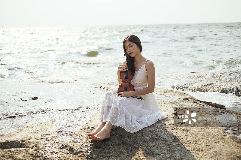 黑色长发白衣女子背靠太阳坐在海边的岩石上拉着小提琴。图片素材