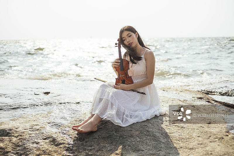 身穿白裙的黑发女子，手拿小提琴，背靠阳光坐在海边的岩石上。图片素材