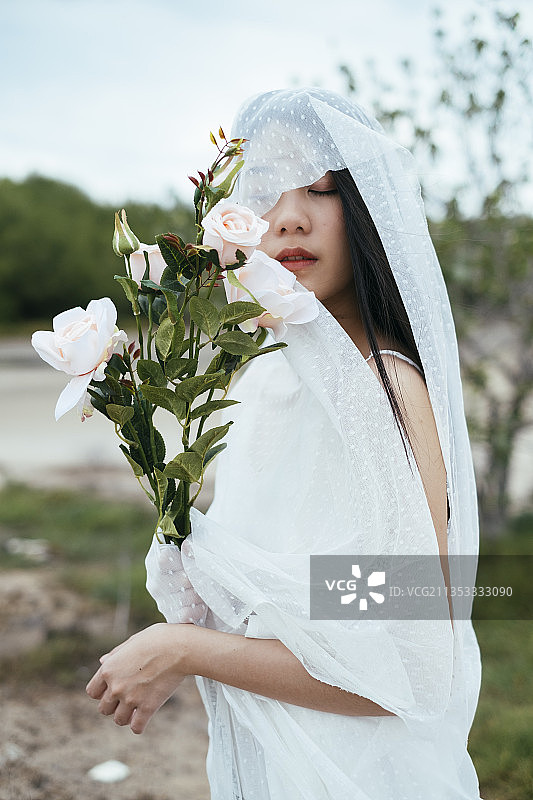特写-闭合的eY年轻的亚洲长黑发妇女新娘与白色面纱举行白色玫瑰花户外。图片素材
