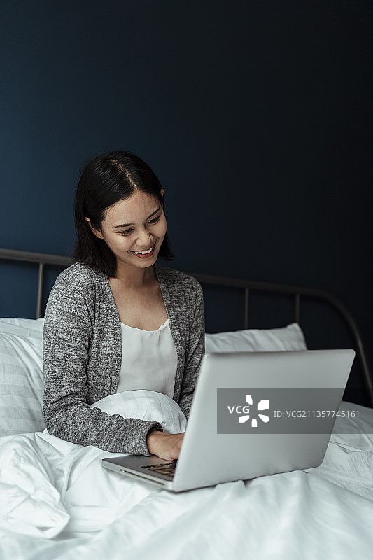 黑色短发女人坐在白色的床上，带着笔记本电脑，微笑着给她的丈夫打视频电话。图片素材