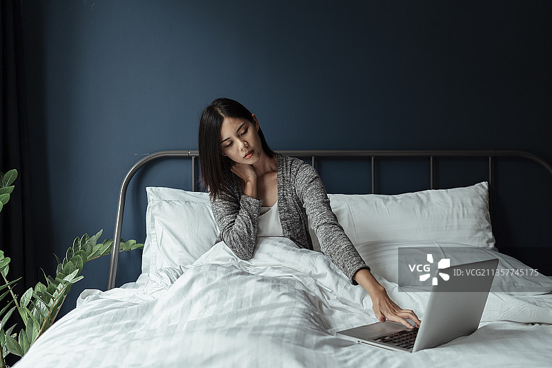黑色短发女人坐在白色的床上，早上用笔记本电脑在她的左边按摩她的脖子。图片素材