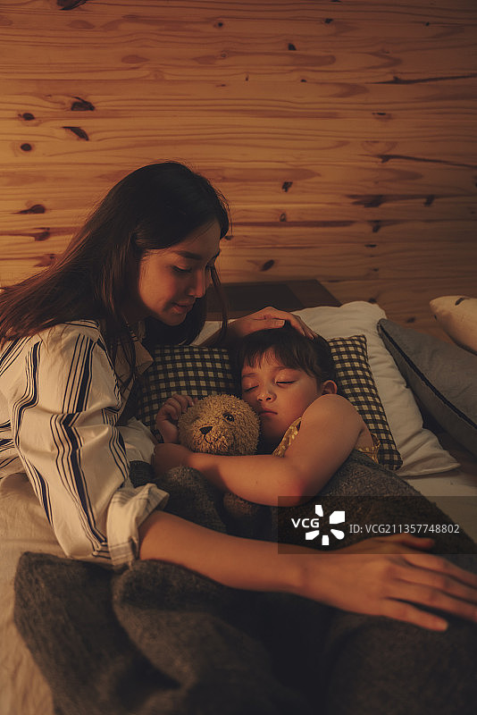 亚洲母亲晚上在床上抱着泰迪熊看她的孩子睡觉。图片素材