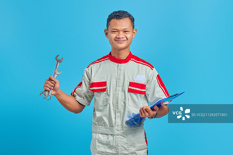 微笑的年轻机械师拿着剪贴板，展示在蓝色背景上孤立的扳手，印度尼西亚图片素材