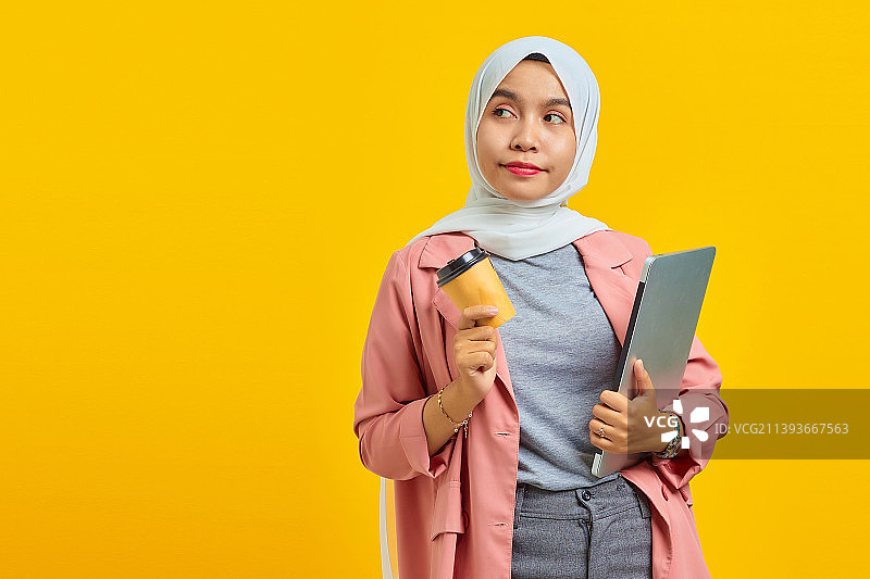 欢快的年轻亚洲女性的肖像，拿着笔记本电脑和咖啡孤立在黄色背景，印度尼西亚图片素材