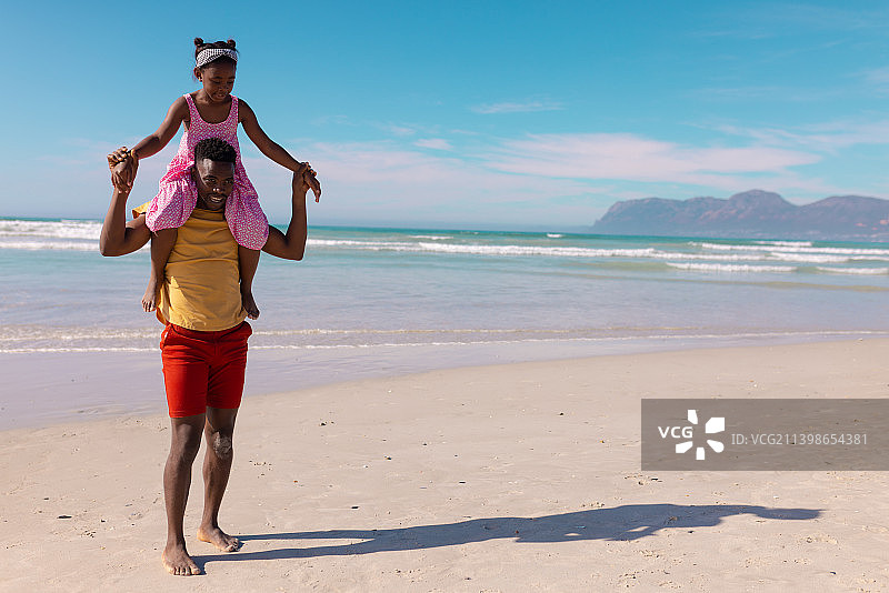 非洲裔美国年轻人背着女儿在海边散步，面对着蓝天图片素材