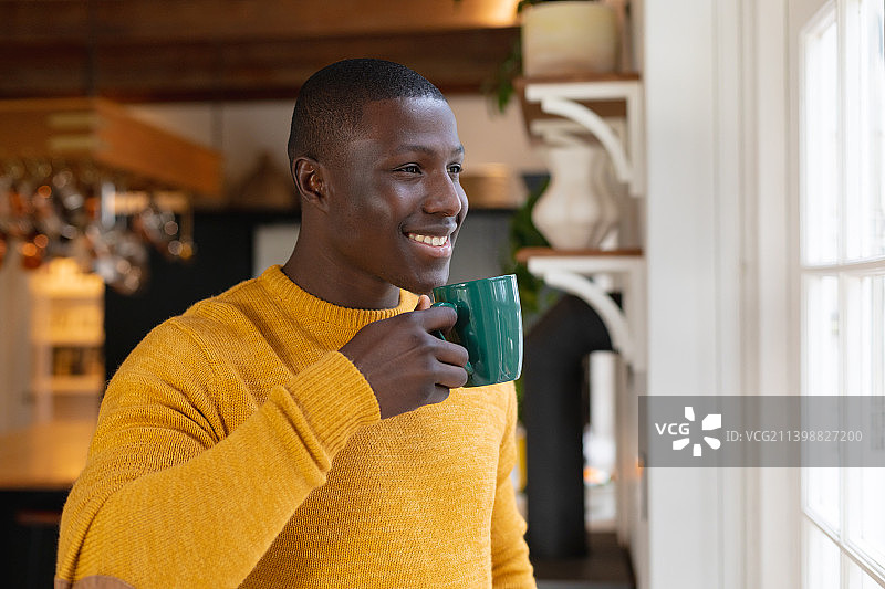 微笑的非洲裔美国人在家里的厨房里喝着咖啡，看着窗外图片素材