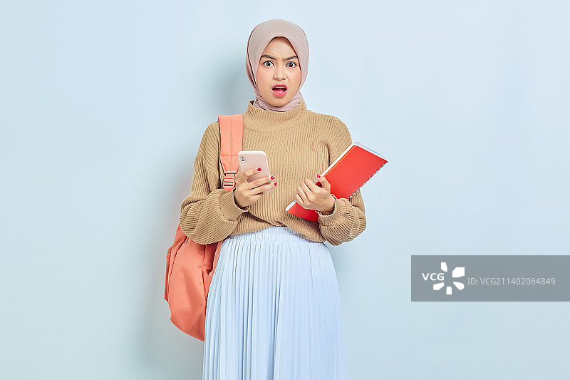 震惊的年轻亚裔穆斯林女学生，棕色毛衣和背包，拿着书和手机图片素材