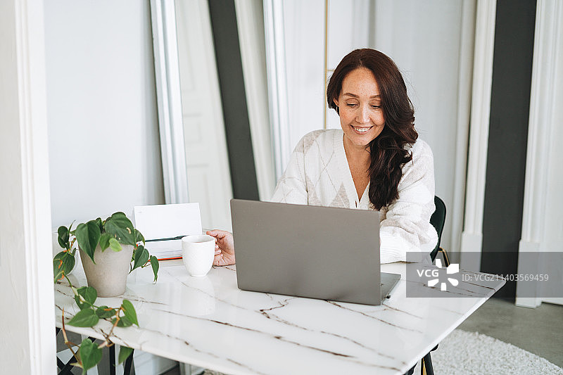 俄罗斯斯维尔德洛夫斯克州叶卡捷琳堡，一名身穿白色开衫的黑发女子，面带微笑，在明亮现代的办公室里用笔记本电脑办公图片素材