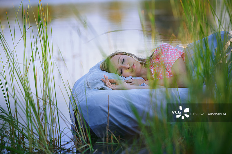 美丽的金发女人穿着粉红色的圆点睡衣睡在漂浮的床在一个湖在日落。很适合做书的封面。超现实主义的概念图片素材