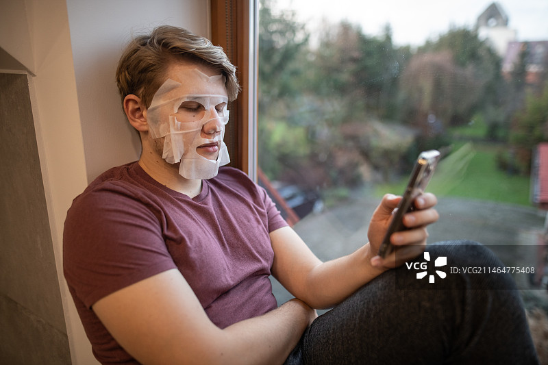 波兰，一名戴着面膜的男子坐在家里的窗台上玩手机图片素材