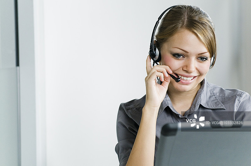 微笑的商务女性戴着耳机，在办公室使用笔记本电脑图片素材