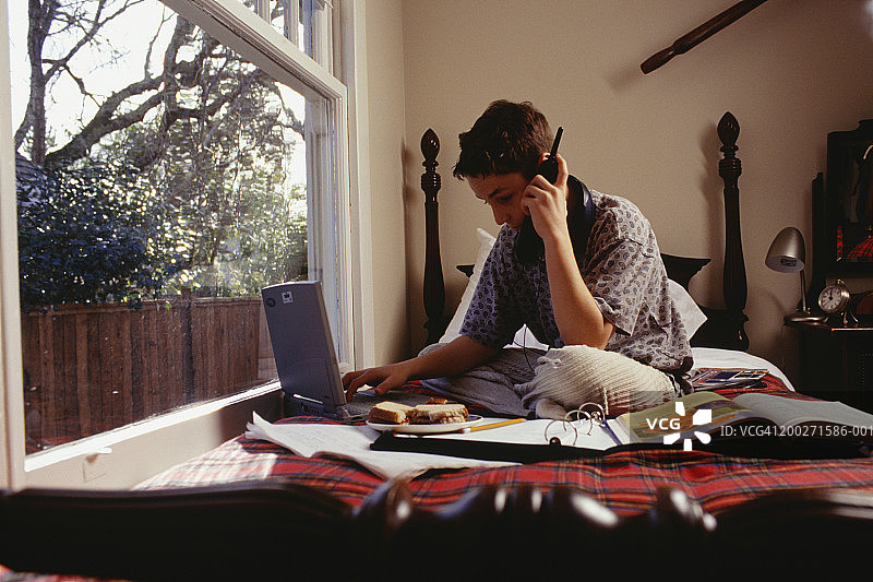青少年男孩(16-17岁)在笔记本电脑上打字和在卧室里使用手机图片素材