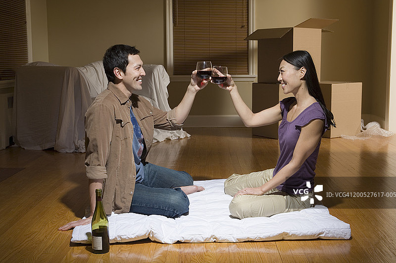 一对夫妇微笑着坐在毛毯上，在没有家具的客厅里，互相敬酒图片素材