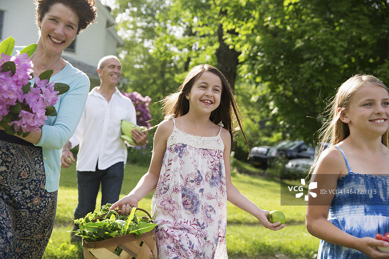 一个夏天在农场的家庭聚会。父母和孩子们带着鲜花、新鲜采摘的蔬菜和水果穿过草坪。为聚会做准备。图片素材