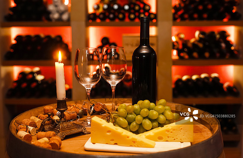 私人酒窖与一瓶红酒，两个酒杯，葡萄，奶酪和点燃的蜡烛在前景图片素材