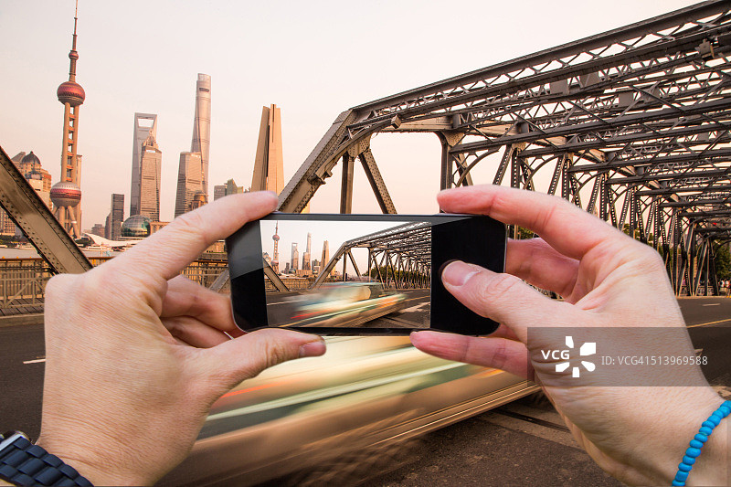 一名男子在外白渡桥上用智能手机从个人角度拍摄上海城市景观，车上有一辆正在行驶的汽车。图片素材