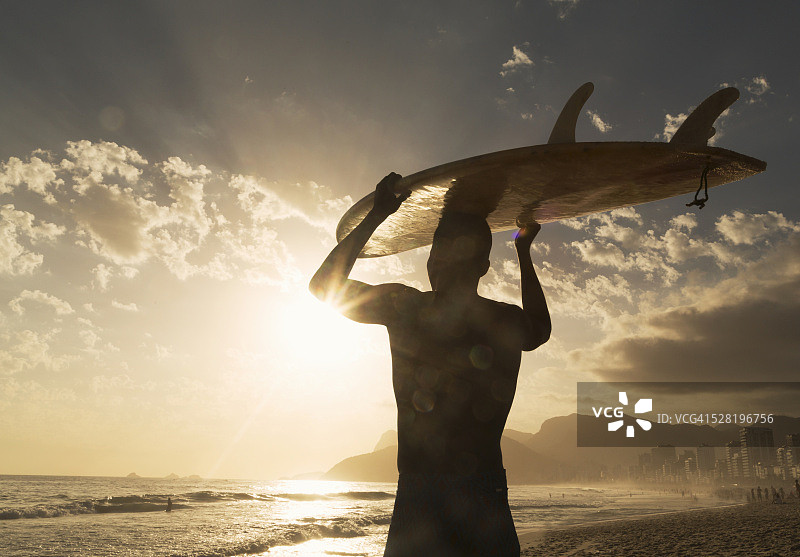 日落时分，在巴西里约热内卢的海滩上，一个冲浪者手持冲浪板图片素材