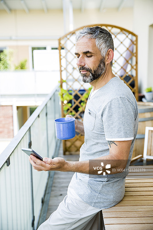 一个男人在阳台上拿着智能手机和一杯咖啡图片素材