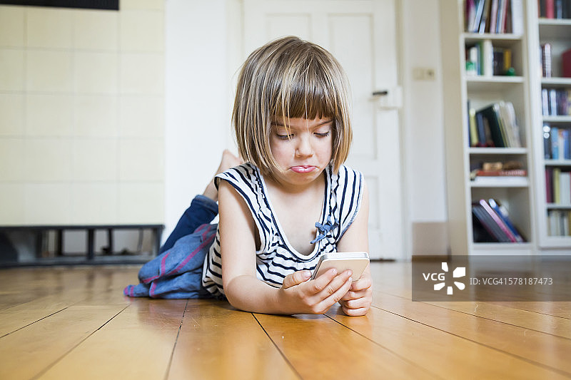 小女孩躺在木地板上，怀疑地看着智能手机图片素材