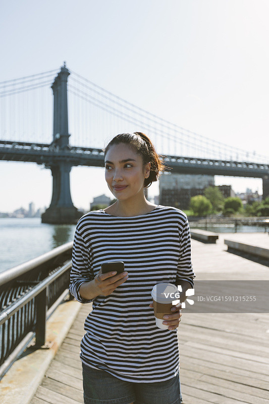 美国，纽约，一幅微笑的年轻女子拿着智能手机和咖啡的照片图片素材