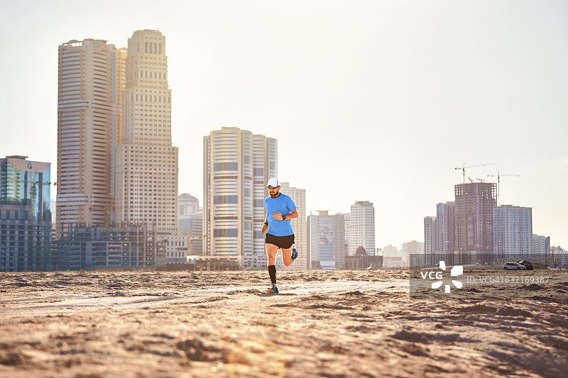 阿拉伯联合酋长国迪拜，一名中年男子在摩天大楼旁的沙地上奔跑图片素材